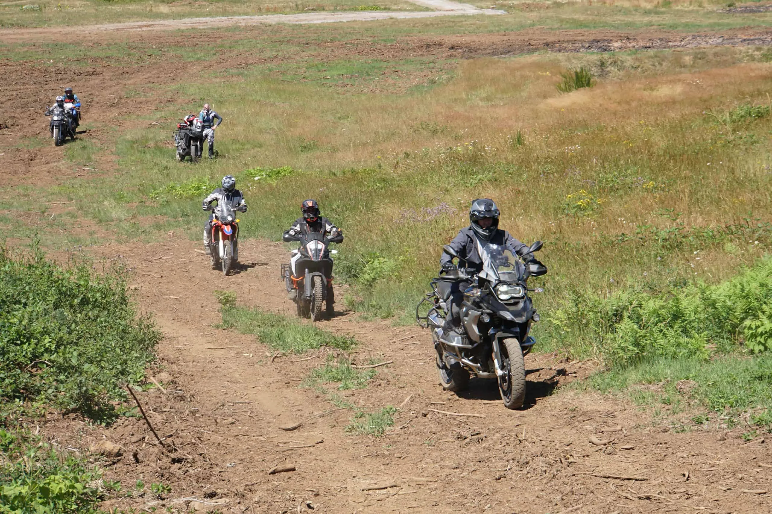 A line of riders on adventure bikes follows a dirt track up a grassy hillside