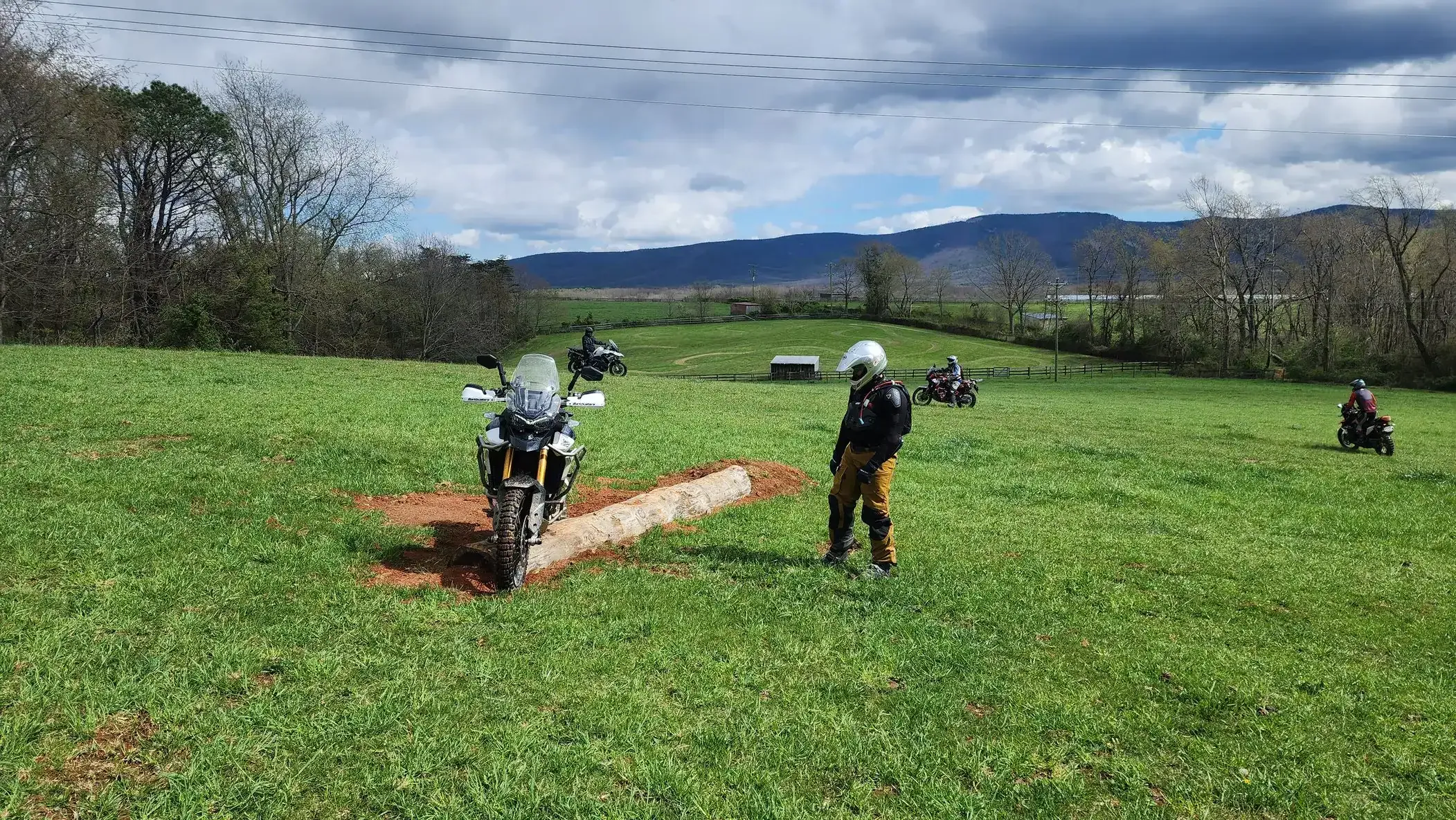 A rider crosses a log obstacle on a grassy field as an instructor watches, mountains behind, Virginia
