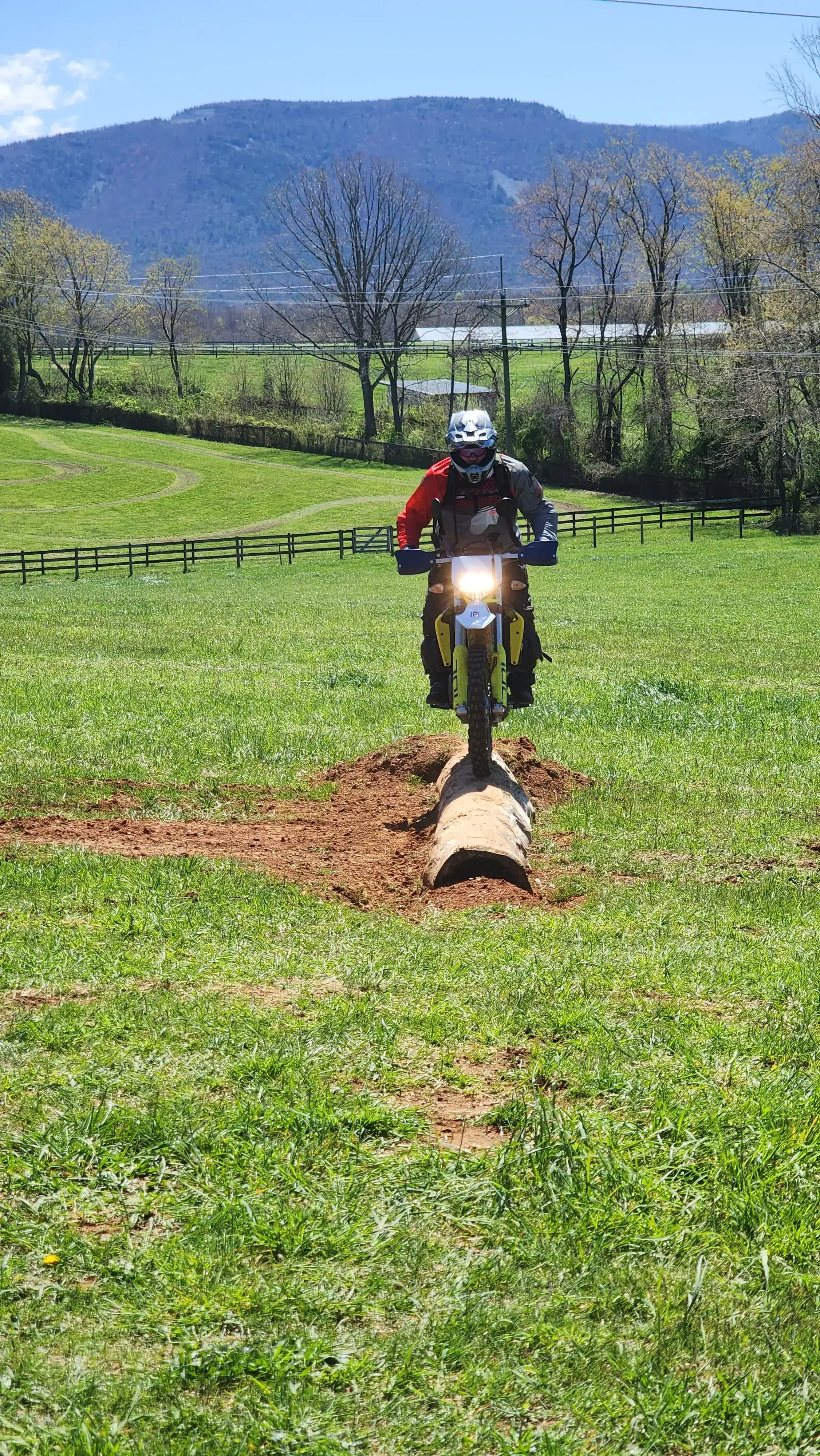 A rider rolls an adventure bike over a log on a grassy field, fences and mountains behind, Virginia