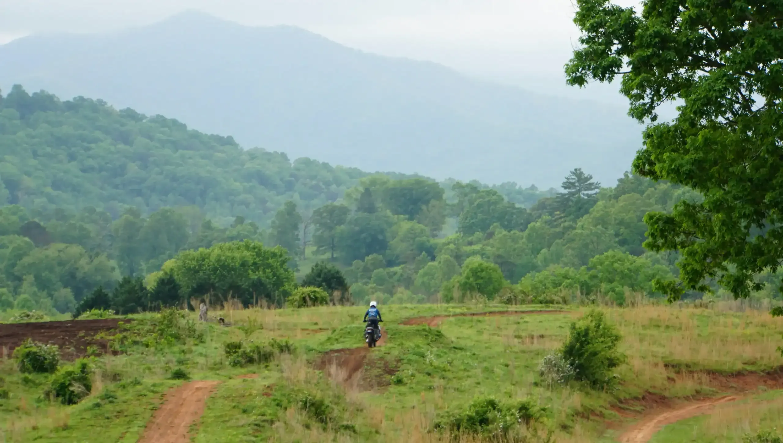 A rider crosses a green hillside on a red-dirt trail below wooded mountains, Georgia