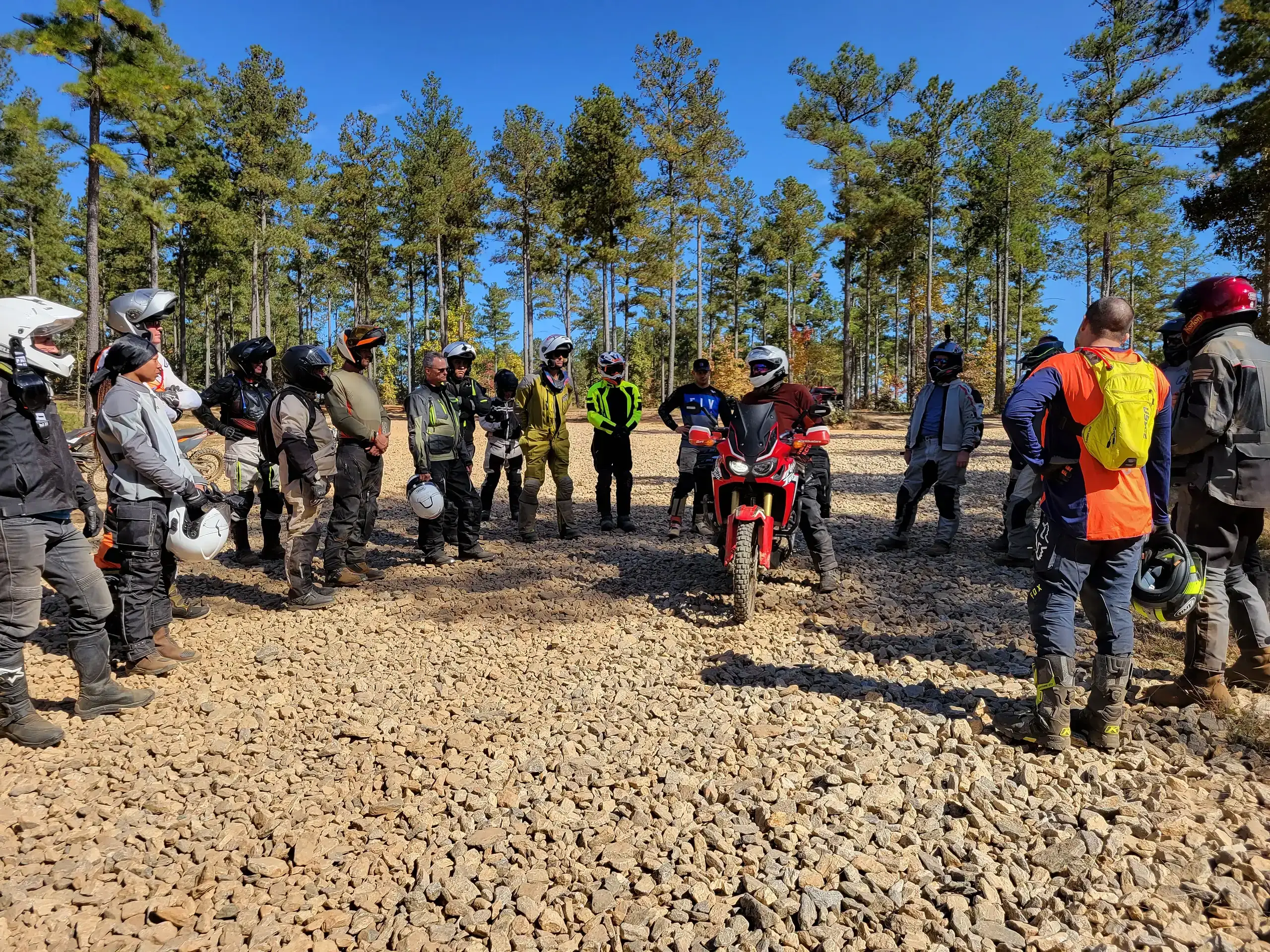 Riders in full gear stand in a circle for a briefing on a gravel clearing among tall pines, Georgia