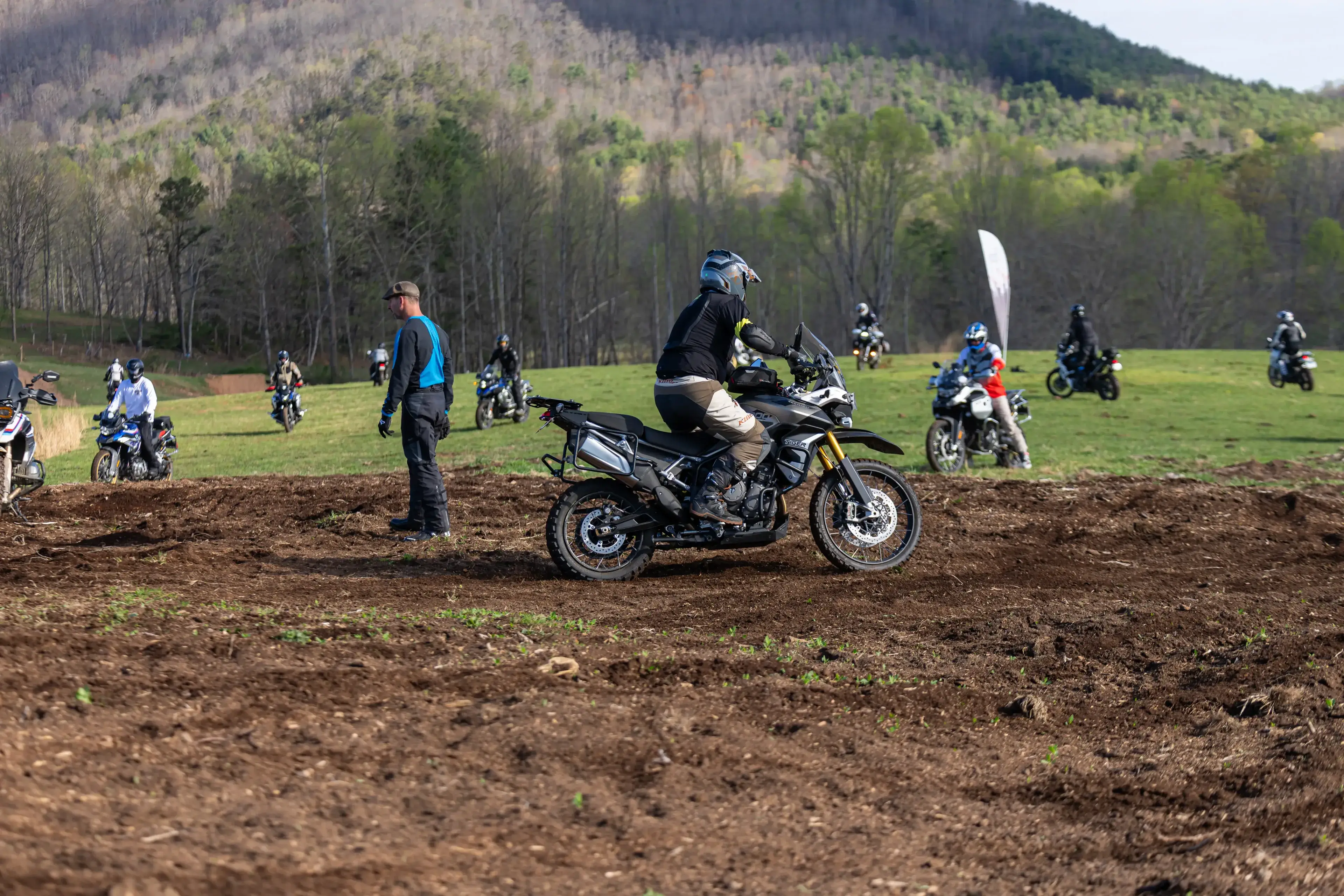 A student rides through a churned mud patch as an instructor watches, riders waiting on the grass, Georgia
