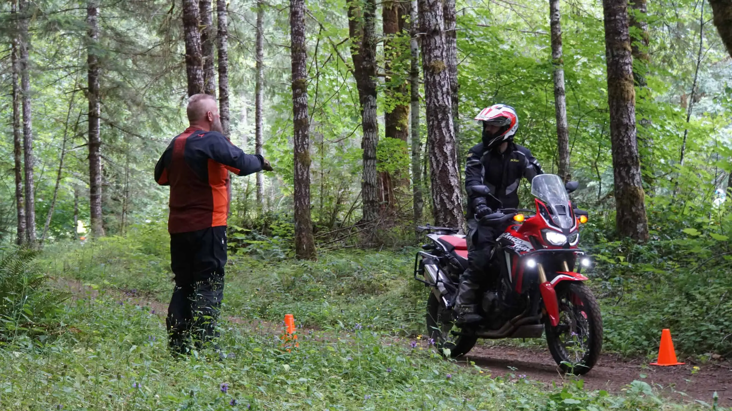 An instructor directs a rider on a red adventure bike between cones on a forest trail