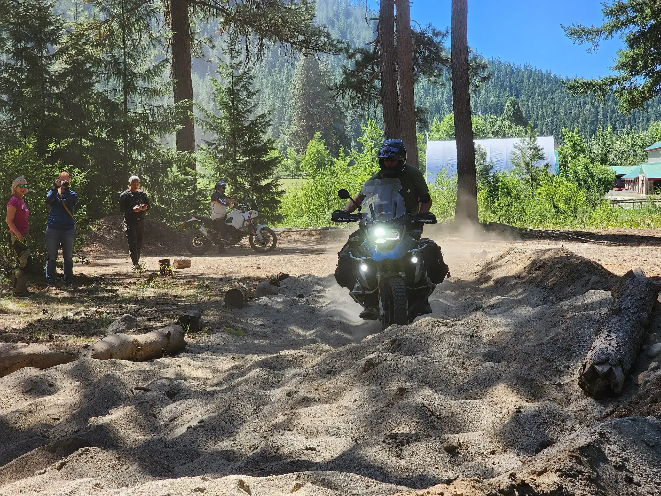 Rider powers a loaded adventure bike through deep sand as others watch, Washington