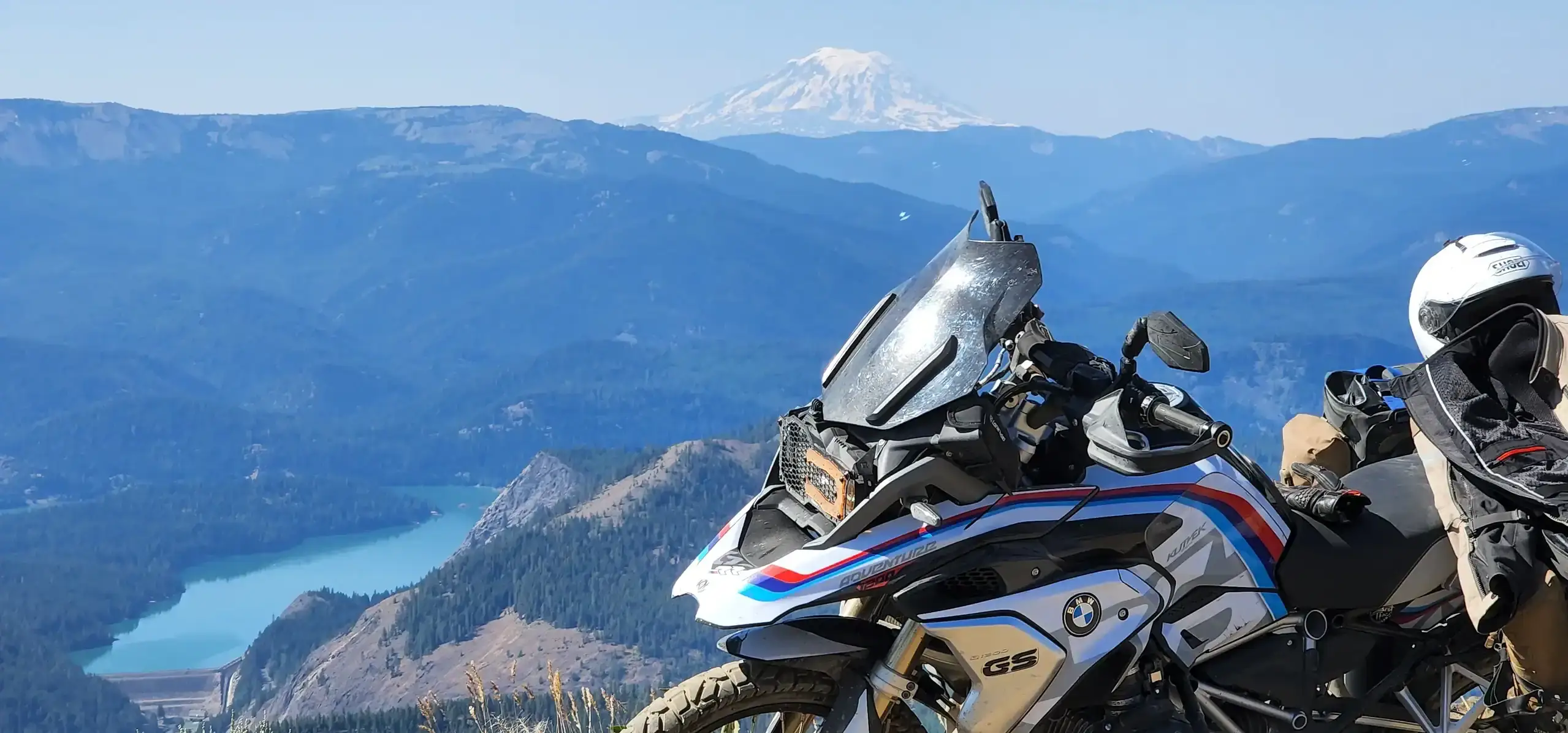 Two adventure bikes stop on a high ridge above a forested valley and a lake, a snowy peak beyond, Washington