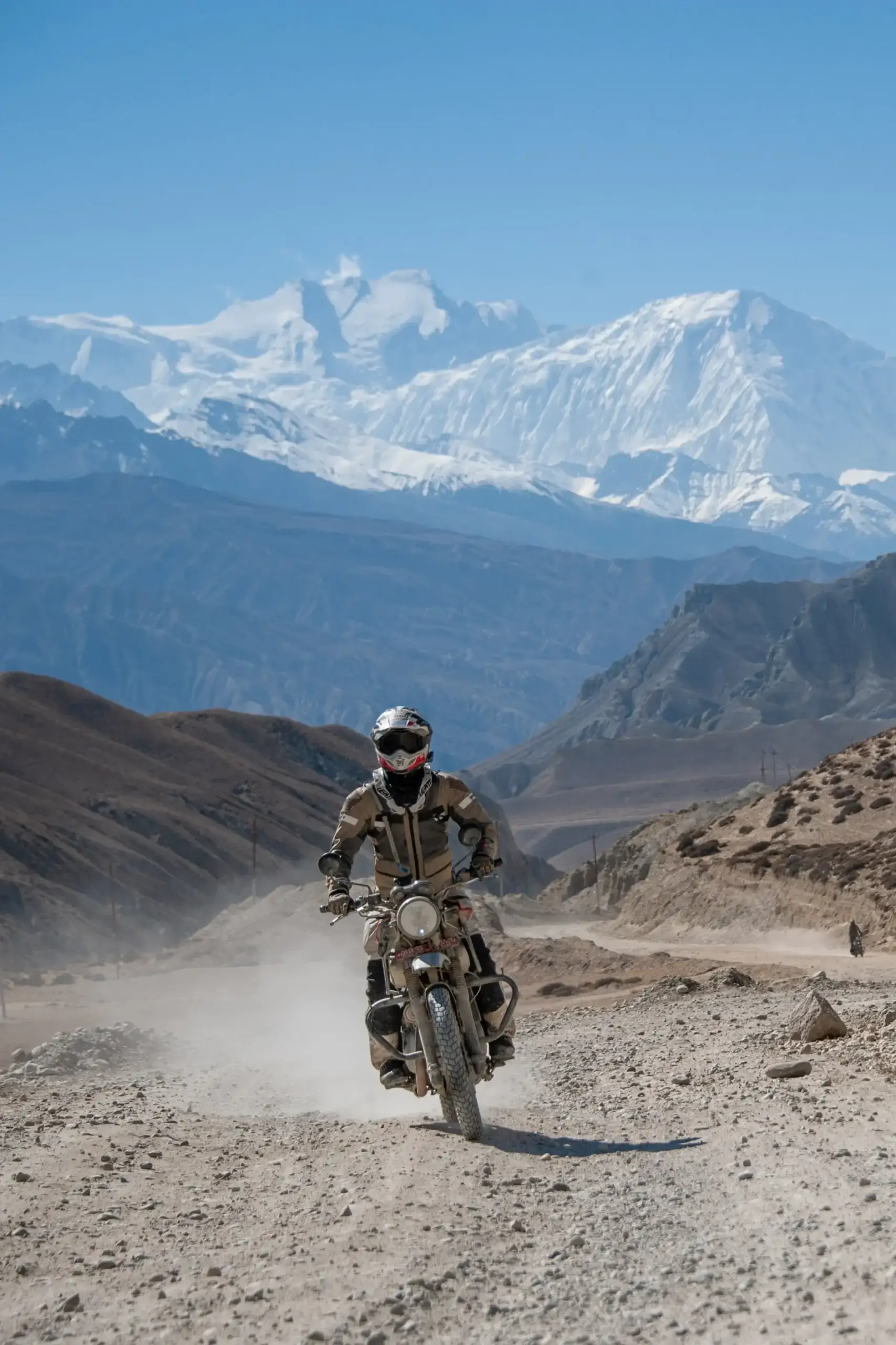 A rider on a Royal Enfield comes down a dusty mountain road below snowy Himalayan peaks, Nepal