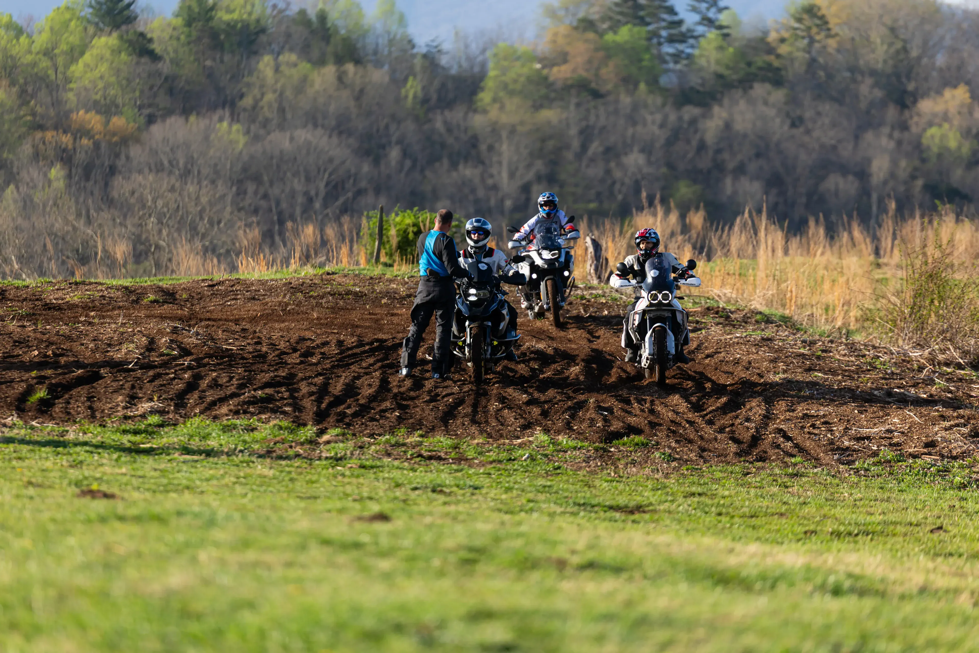 Bret Tkacs coaches riders on adventure bikes through a churned mud patch, wooded hills behind, Georgia