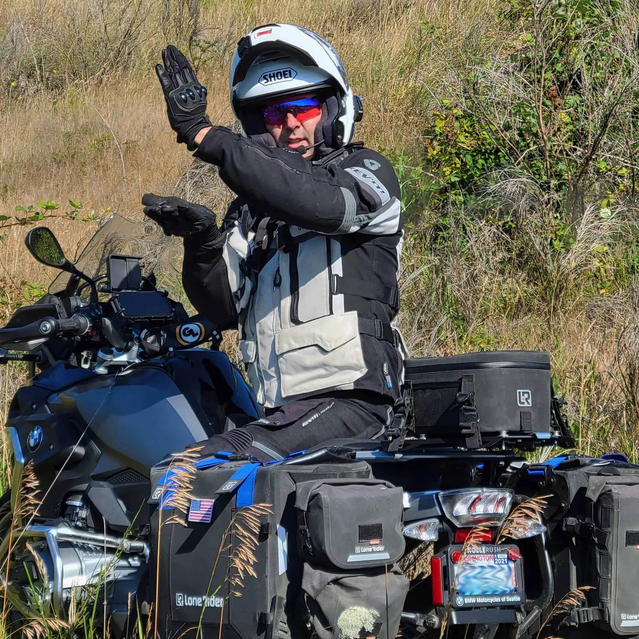 A rider in full adventure gear signals from a loaded BMW adventure bike on a grassy hillside